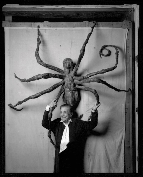 In a black and white photograph, the artist Louise Bourgeois reaches overhead and holds the legs of a large spider sculpture overhead.