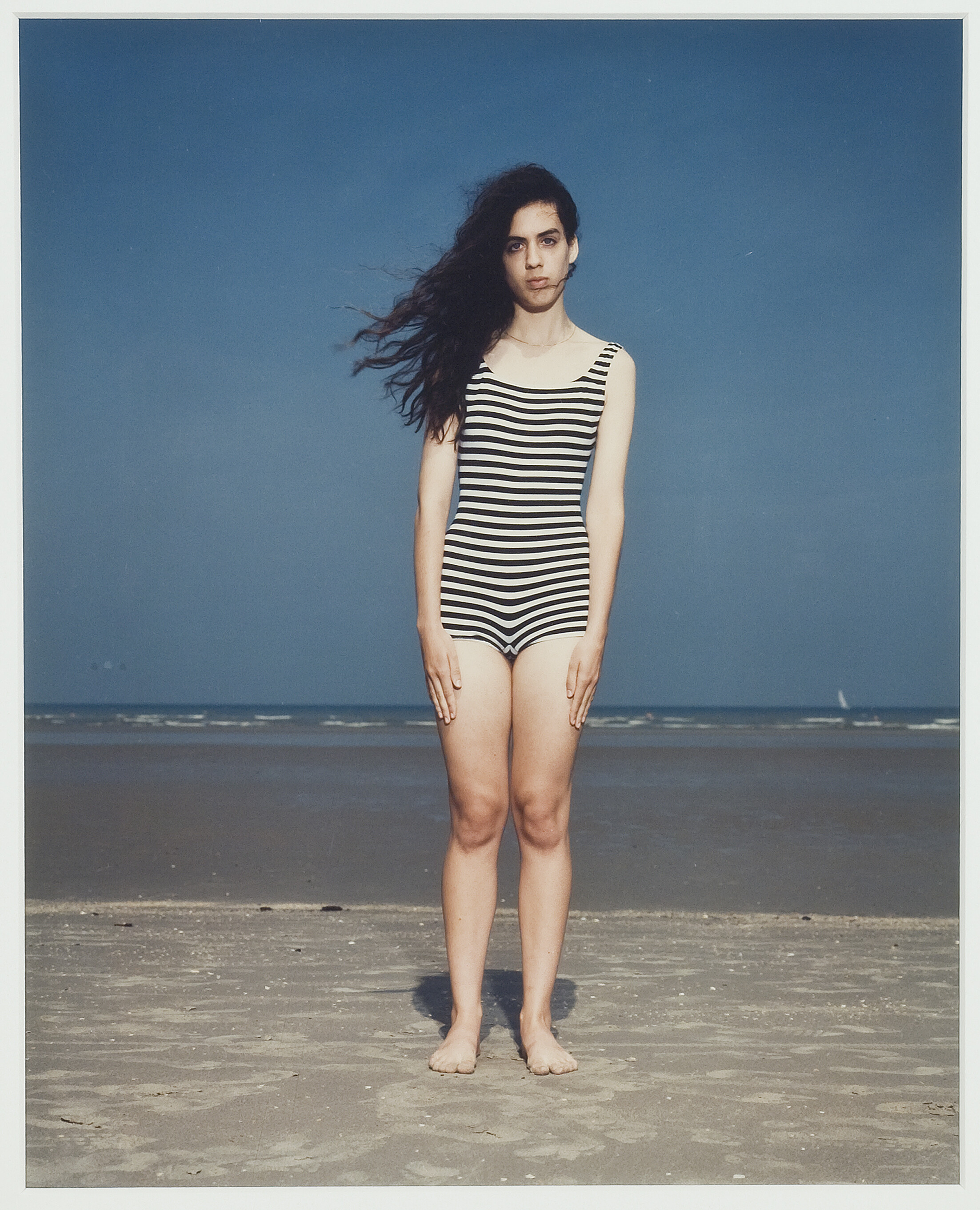 A light-skinned person wearing a black and white striped bathing suit standing at the beach with their back turned towards the ocean. Their long dark hair is blowing in the wind and their arms are held straight at their sides.