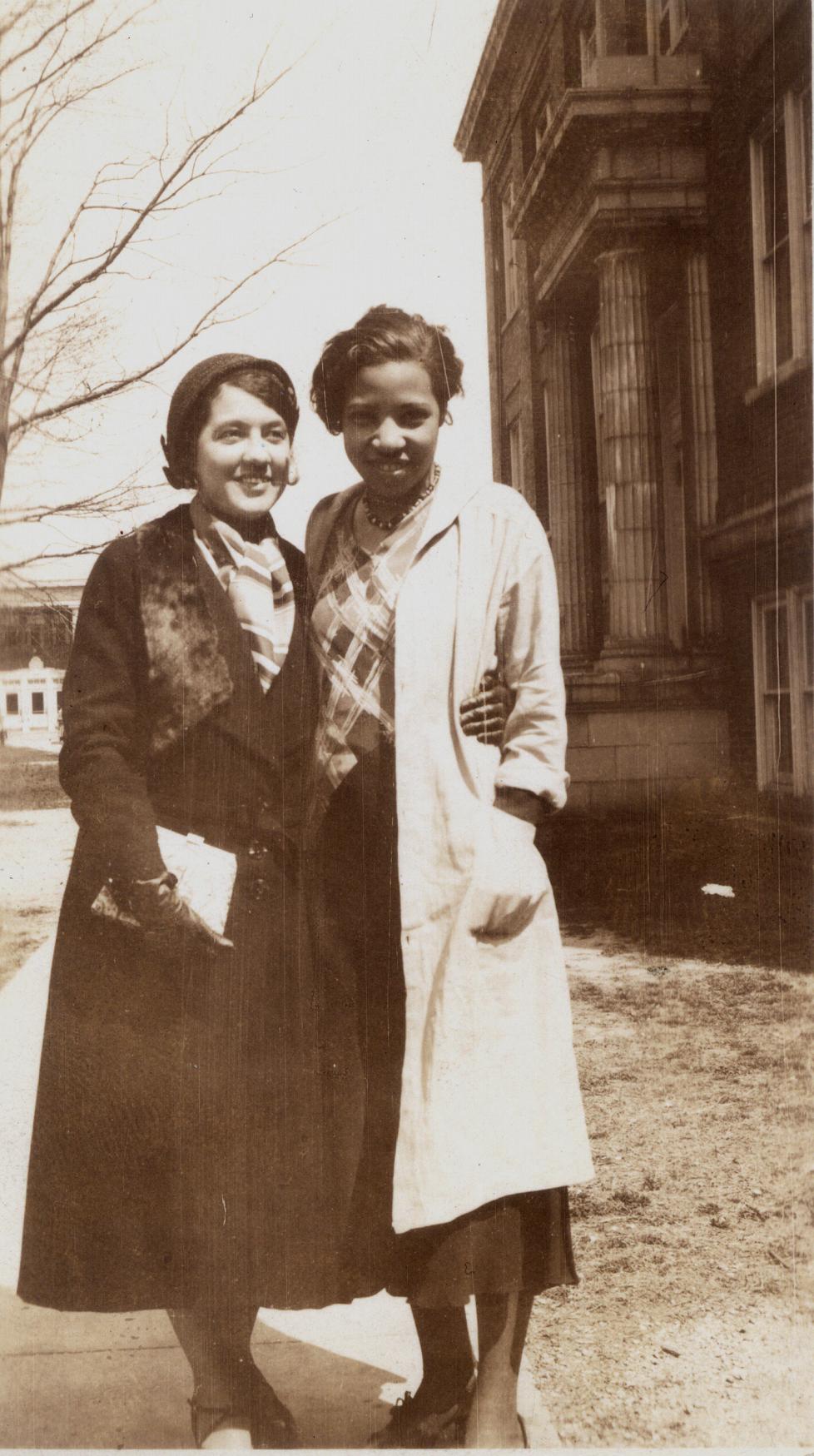 A sepia-toned photograph of two women standing with their arms around each other. They are both wearing long coats and dark skirts.