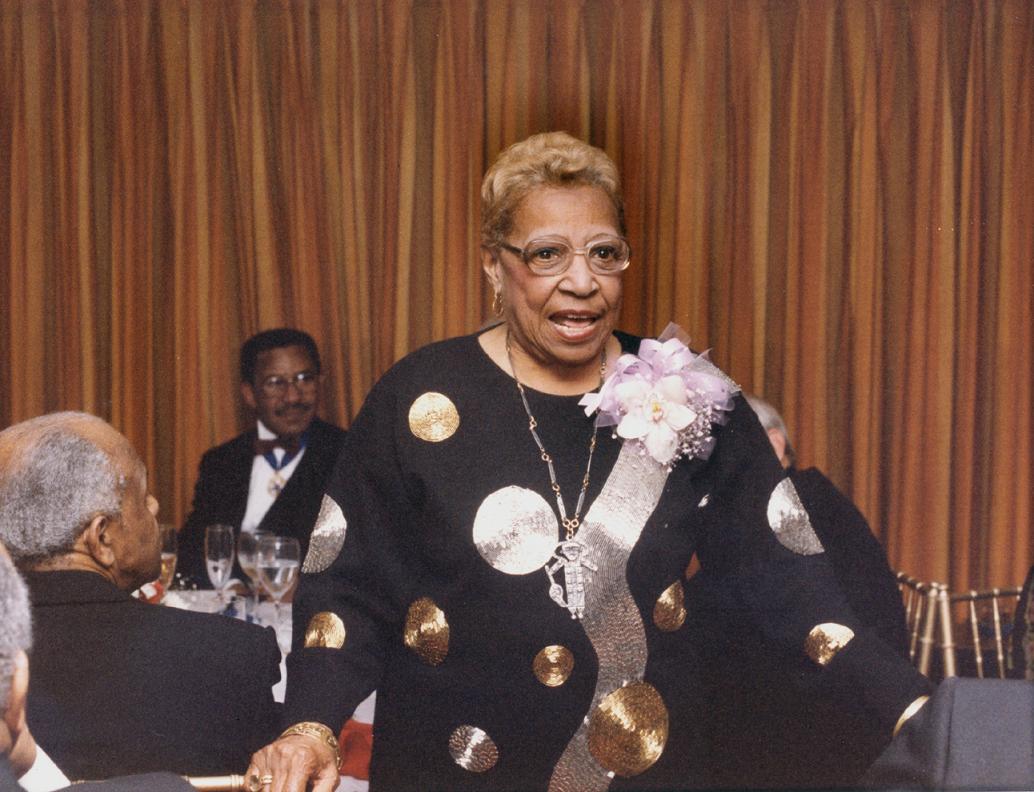 A medium-skinned African American woman wearing glasses and a black dress with metallic polka dots. She is in the middle of speaking while people wearing suits seated in the background listen.