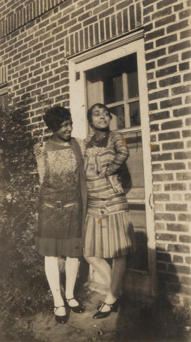 A black and white photograph of two medium-skinned African American women wearing 1920s style dresses, white stockings, and black heeled shoes standing in front of a door.