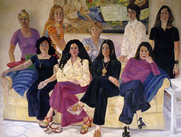 Five women stand against a yellow wall and in front of them four more women pose cross legged on a couch. 