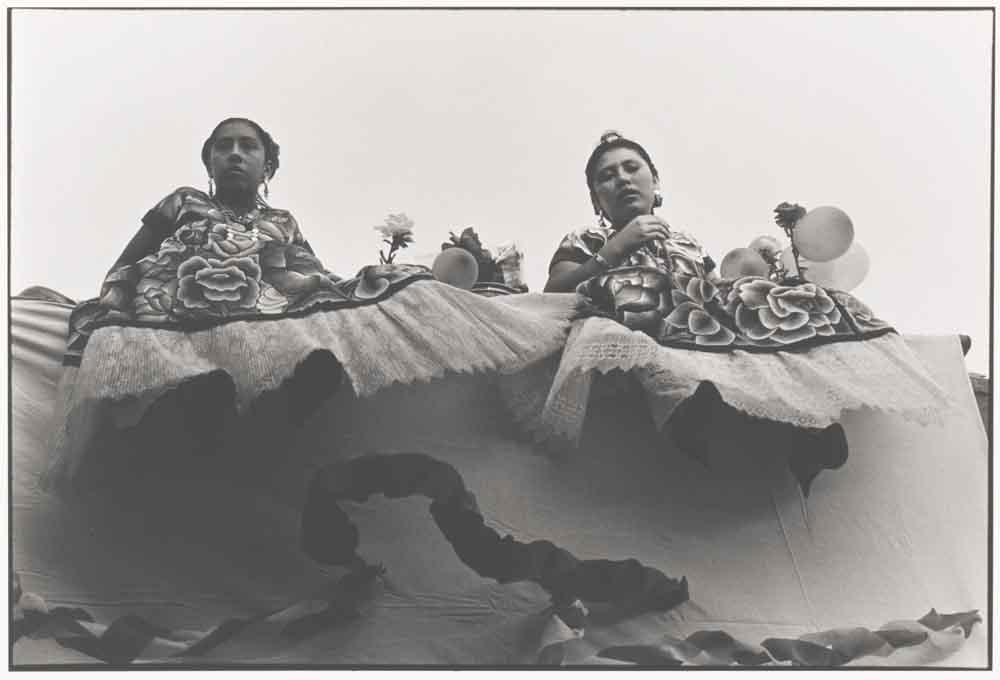 Two women in traditional Mexican dresses sitting on a wall photographed from below.