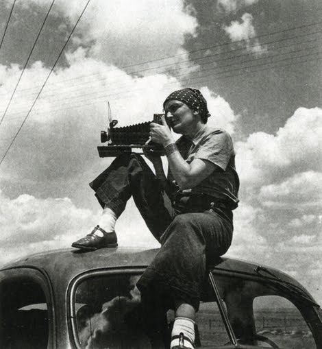 A woman with a light skin tone sitting on the top of a car with a camera.