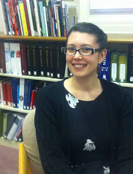 A woman with a light skin tone and black hair sitting before a shelf with books.