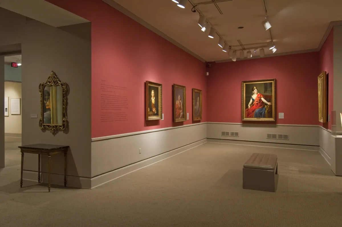 View of a gallery space with red walls. A golden mirror with plenty of ornaments is hanging to the left, large historical portraits of women are hanging to the right.