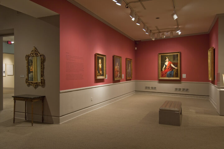 View of a gallery space with red walls. A golden mirror with plenty of ornaments is hanging to the left, large historical portraits of women are hanging to the right.