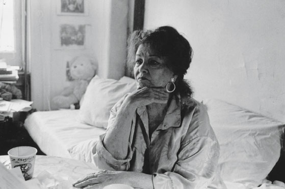 A black-and-white photograph of a woman sitting by a bed.