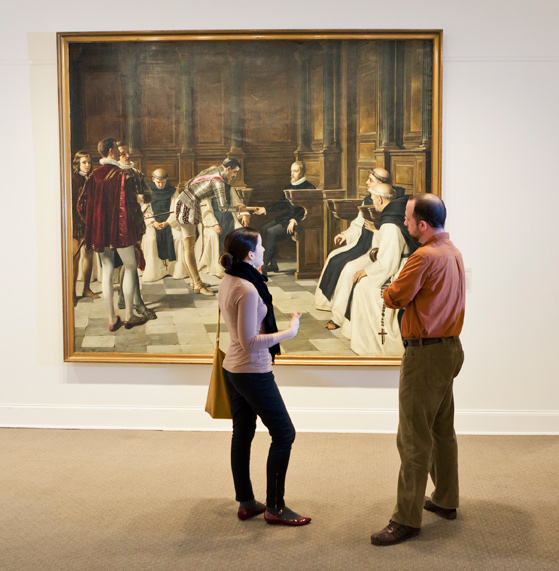 Two museum visitors are standing before a large oil painting, discussing it.