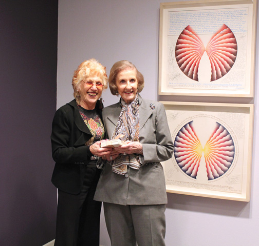 Two women with a light skin tone and gray and blonde hair are posing in front of a colorful artwork.