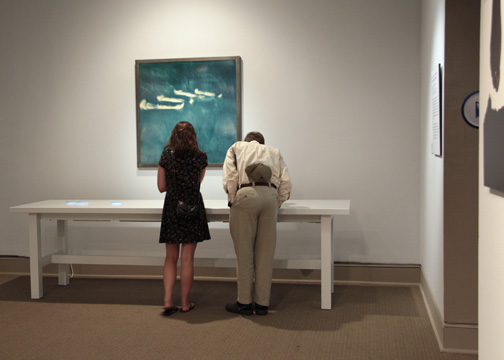 Two people looking at artwork on a white table in a white gallery space; behind the table is a navy artwork with four thick, white lines on it.
