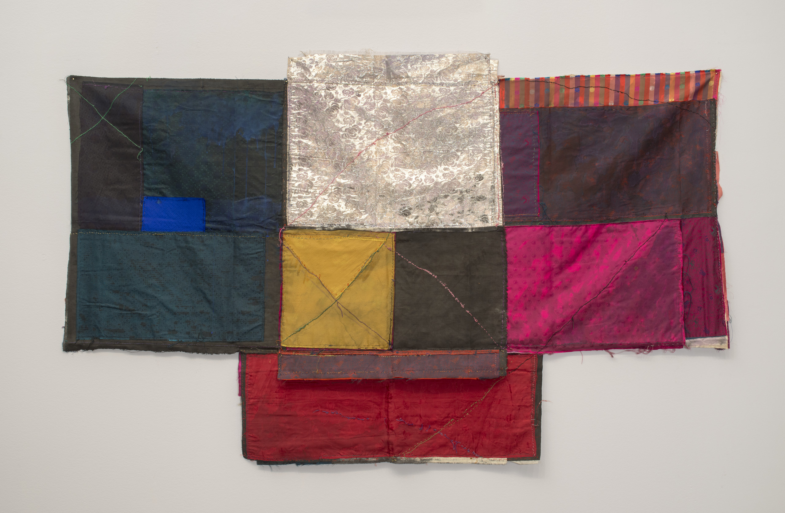 Textile collage of cloth folded and stitched together in loose geometric formations with visible seams making linear patterns. Deep blues on the left and warm colors on the right frame blocks of silver, yellow and grey in the center, which extends into a slightly longer base.