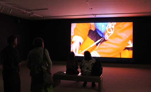 Four audiences sitting in a dark room watching Kimsooja's Thread Routes-Chapter 1. On the screen there is a pair of arms from a person wearing a red jacket and a blue shirt.