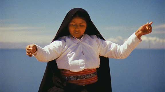A medium skin-toned adult woman raises both her arms in front of a blue sky, holding thread. She has a long, black cloth covering her hair and draping behind her like a cloak, and she wears a white long-sleeved shirt and a red belt.
