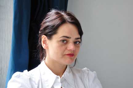 Portrait photograph of a woman with a light skin tone and black hair in a bun, wearing a white shirt and red lipstick.