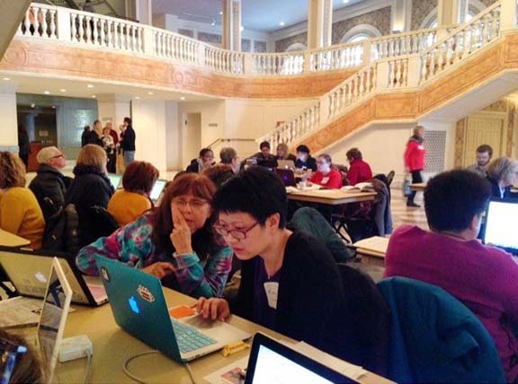 Several groups of people are sitting in a large foyer around tables on laptops. Two women on a laptop are in focus.