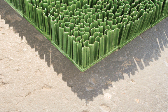 Detail photograph of ceramic sculpture made to look like a patch of lawn. Individual squares consisting of multiple upright blades of porcelain grass, glazed green, fit together to form a lush rectangular field of grass.