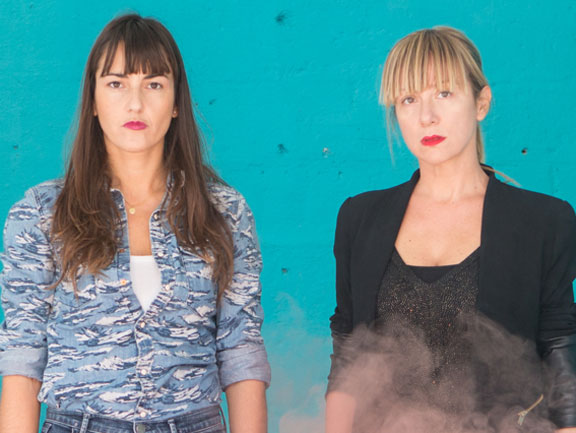 Portrait photograph of two women standing in front of a blue wall. They have light skin tones and long brown as well as short blonde hair.