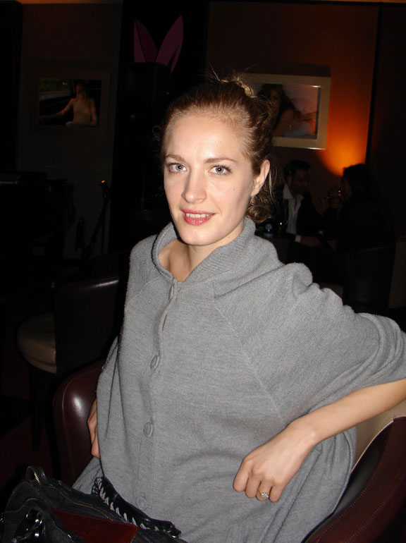 Polly Morgan sitting and smiling in a dark room, seen from the waist up. She is a light-skinned adult woman with blonde hair pulled back in a bun, wearing light makeup and poncho-like, button-up, gray top.