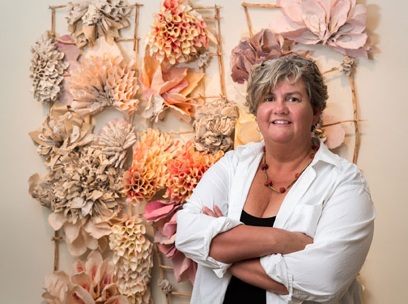 A woman with a light skin tone and short blonde hair standing in front of flowers made out of paper assembled on a wall.