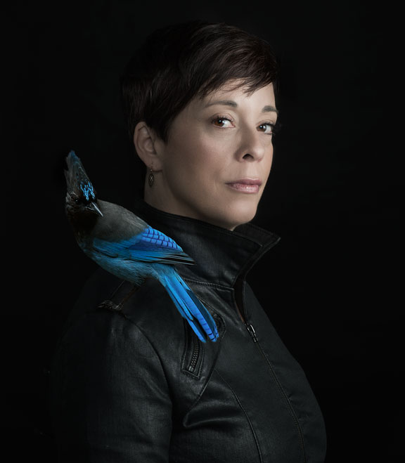 A portrait photograph of a woman with a light skin tone and a black and blue bird sitting on her shoulder.