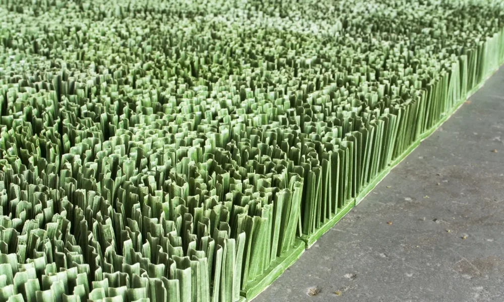 Detail photograph of ceramic sculpture made to look like a patch of lawn. Individual squares consisting of multiple upright blades of porcelain grass, glazed green, fit together to form a lush rectangular field of grass.