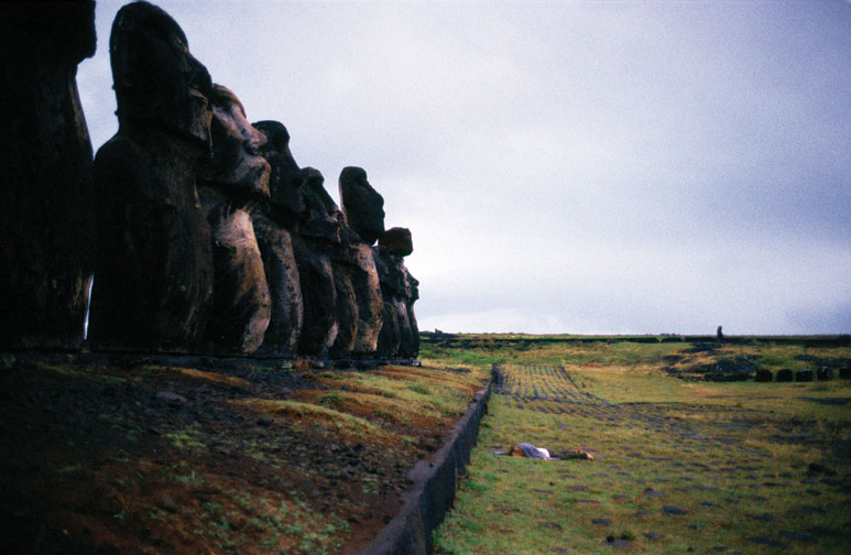 A color photo of the Moai, monolithic human figures carved by the Rapa Nui people on Easter Island. The statues are arranged in a line to the left. Before them, a person laying face down on the grass with their limbs sprawled out.