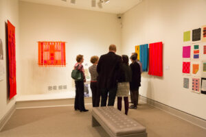 View of a gallery space. Several people are standing in a circle, discussing the artworks on the wall. The artworks are colorful textile art pieces.