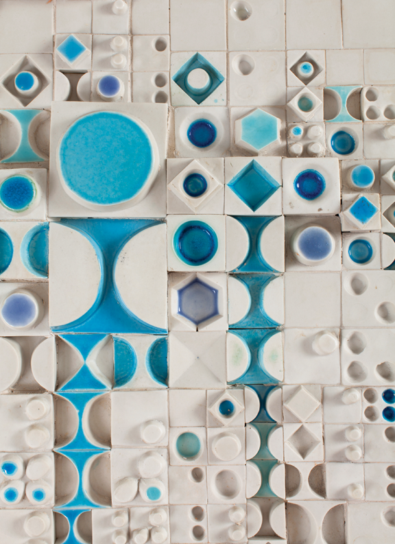 A wall of white ceramic tiles with dark blue und light blue geometric shapes cut into them. The cut-outs create a haptic, 3d-effect.