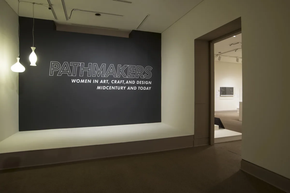 View of a gallery space. On a black wall, it says " Pathmakers: Women in Art, Craft, and Design, Midcentury and Today" in big, white letters, Two pendant lights are hanging from the ceiling to the left.