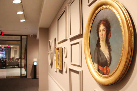 View of a gallery space with several paintings in golden frames, as well as white, empty frames. A portrait of a woman with a light skin tone and wavy, brown hair. She is wearing an olive green dress with a red bow around her waist, sitting before a blue background that is reminiscent of a blue sky. She has bright red cheeks and looks towards the viewer with a steady gaze.