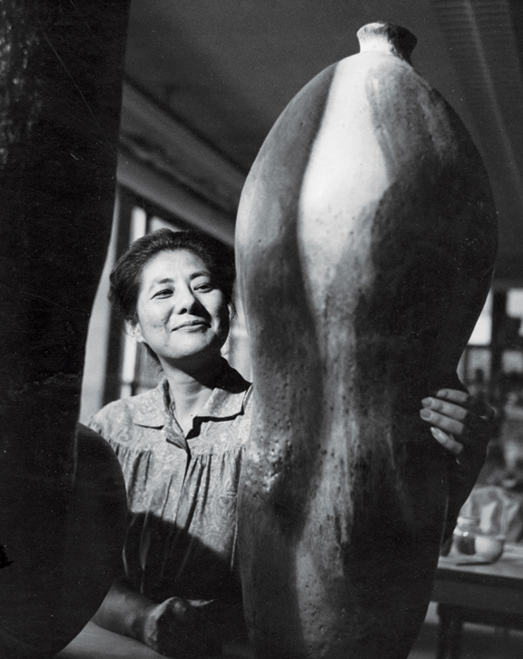 A black-and-white photograph of a light-skinned, dark-haired, Asian adult woman smiling while wearing a smock in her studio. In the foreground are two of her large sculptures; she has her hand on one of them.