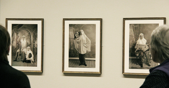 Two museum visitors are viewing three photographs of women in headscarfs posing with a boom box.
