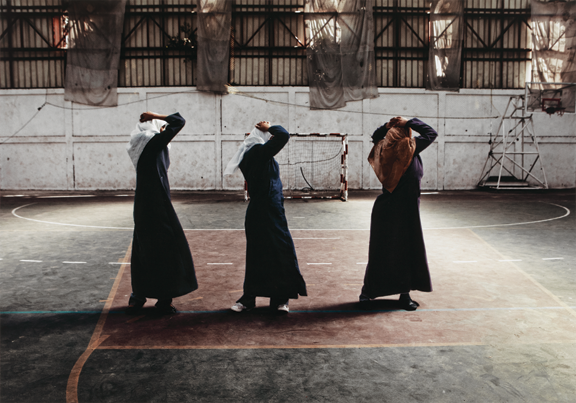 Three women in long coats and headscarfs are dancing in a warehouse building.