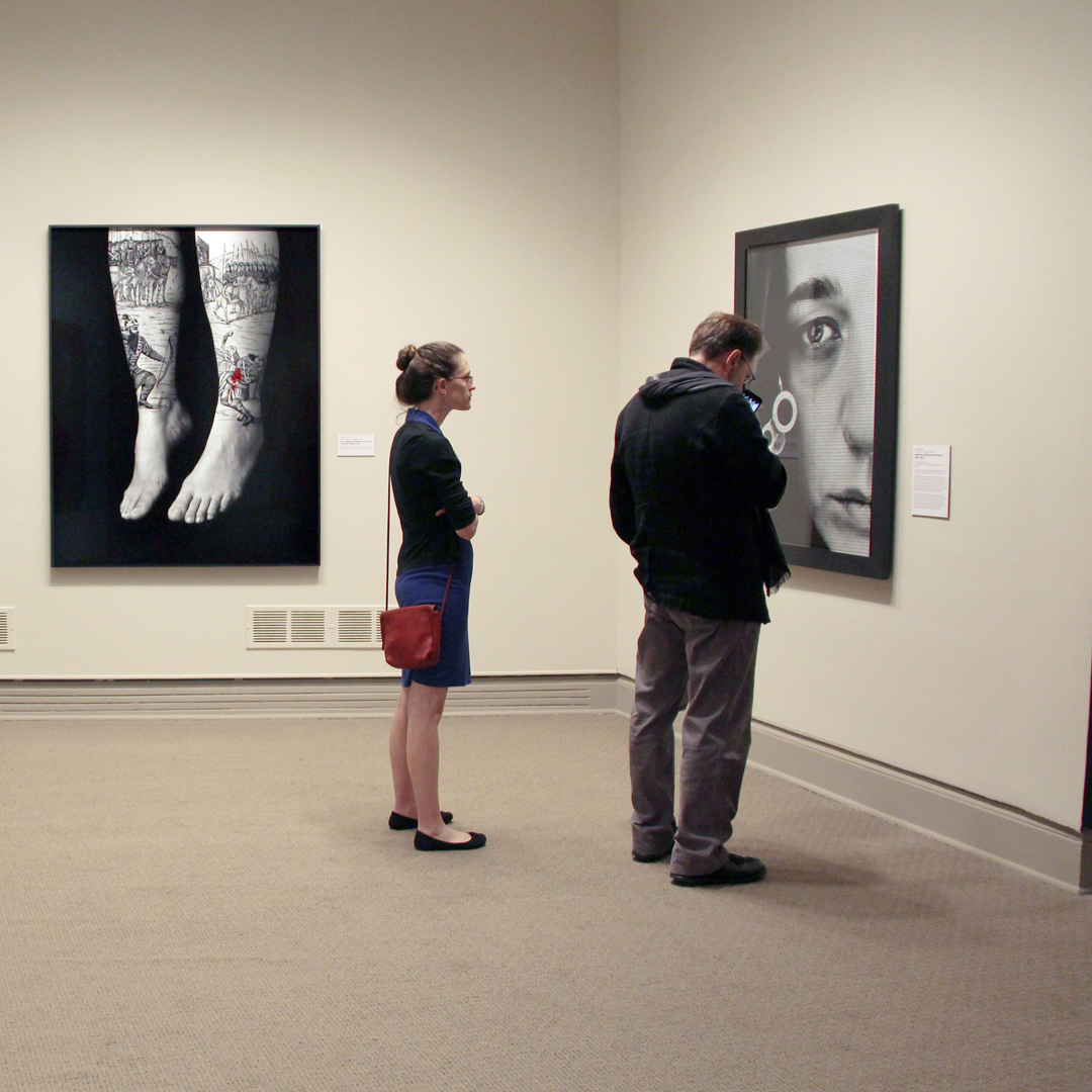 A man and a woman with a light skin tone are standing in a gallery. They are both looking at a large black and white photograph of a woman.
