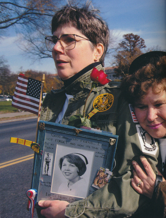 A light-skinned adult with short brown hair and glasses wears a green U.S. Army jacket decorated with patches, and holds a framed, decorated, black-and-white portrait of a nurse who served in the military. The person stands beside a road and another light-skinned older adult hugs them from behind.