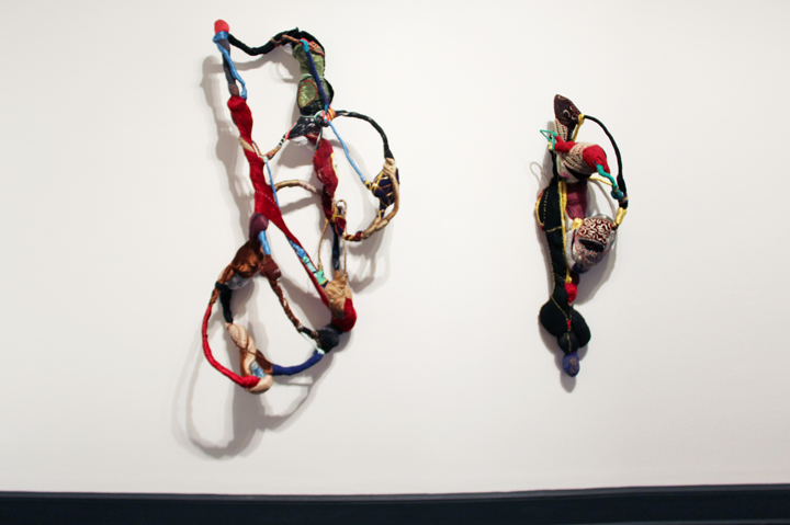 Sculptures made of colorful textiles are hanging on a wall. The texture seems to be either felt or knitted. The sculptures have a delicate look to them, almost as if they were skeletons.