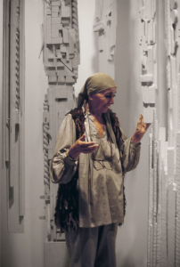A photograph of a light-skinned woman wearing a a olive green headwrap looking at a white totemic wooden sculpture. 