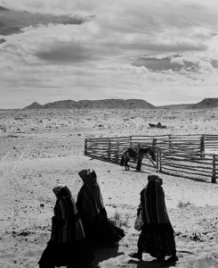 A black and white photograph of three women in long, black dresses and with head coverings walking in a desert landscape. There are mesas in the background, as well as a fence and a horse.