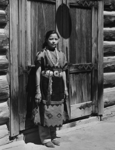 A black and white photograph of a girl in traditional Navaho clothing. The girl has pigtails and is standing before a large, wooden door. 