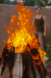 A woman with a light skin tone and short gray hair is standing before a big blazing fire. She is looking directly into the fire. Behind her is a red wall in the style of pueblo architecture.