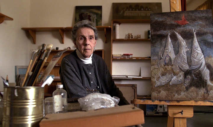 A light-skinned woman with grey hair sitting in an artist studio with paint brushes on a table in front of her and a painting of hooded figures behind her.