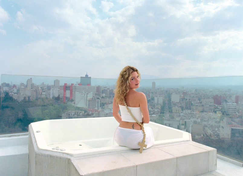 A blonde woman in her twenties in a white tube top and a sheer white skirt perches on the edge of a hot tub and looks over her right shoulder. Around her shoulder hanging diagonally is a large wooden rosary. In the background, a cityscape is set against a blue cloudy sky.