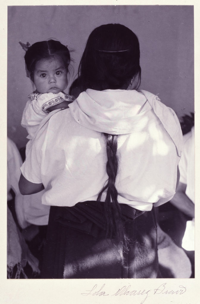 A woman is photographed from behind. Her long black braid trails down the center of her back, and she holds a young female child. The child looks directly at the camera from over the woman’s left shoulder. The sleeves and collar of the child’s white dress have floral borders.