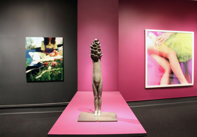 Installation view of a gallery space featuring a sculpture prominently in the middle. The sculpture made of stone resembles a figure, yet instead of a head, there is a pine cone. The figure does not have arms either, it is a biomorphic being reminiscent of fairytale creatures.