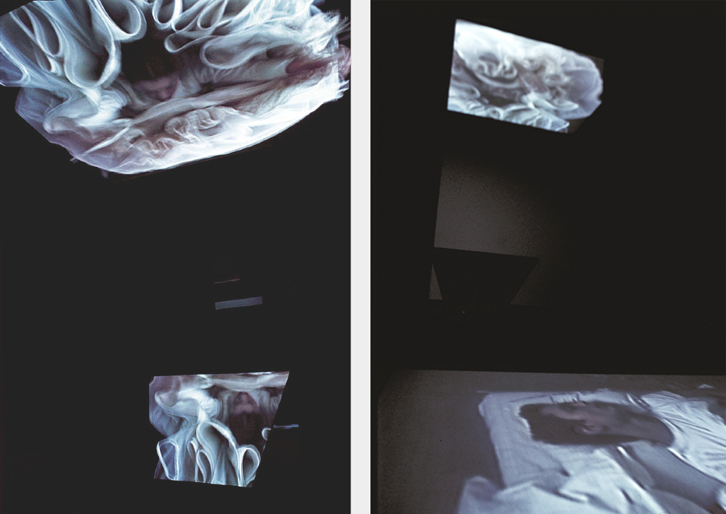 An installation view of photographs projected onto screens in a dark gallery space. On the left, two large photos show a woman lying amidst her big puffy white tulle skirt, with only her head and hands sticking out. In the right image, a photograph of a man lying on a pillow is projected onto a space on the floor.