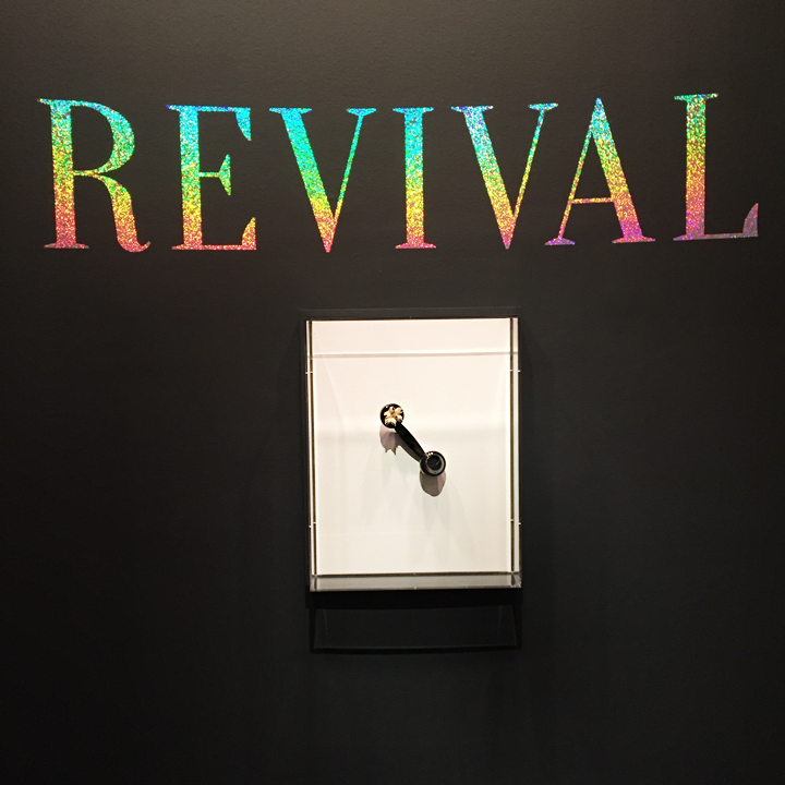 On a black wall, big letters in glitter say "Revival". Underneath is a glass box that showcases a telephone receiver. Five little chicken are exiting from the upper part of the receiver.
