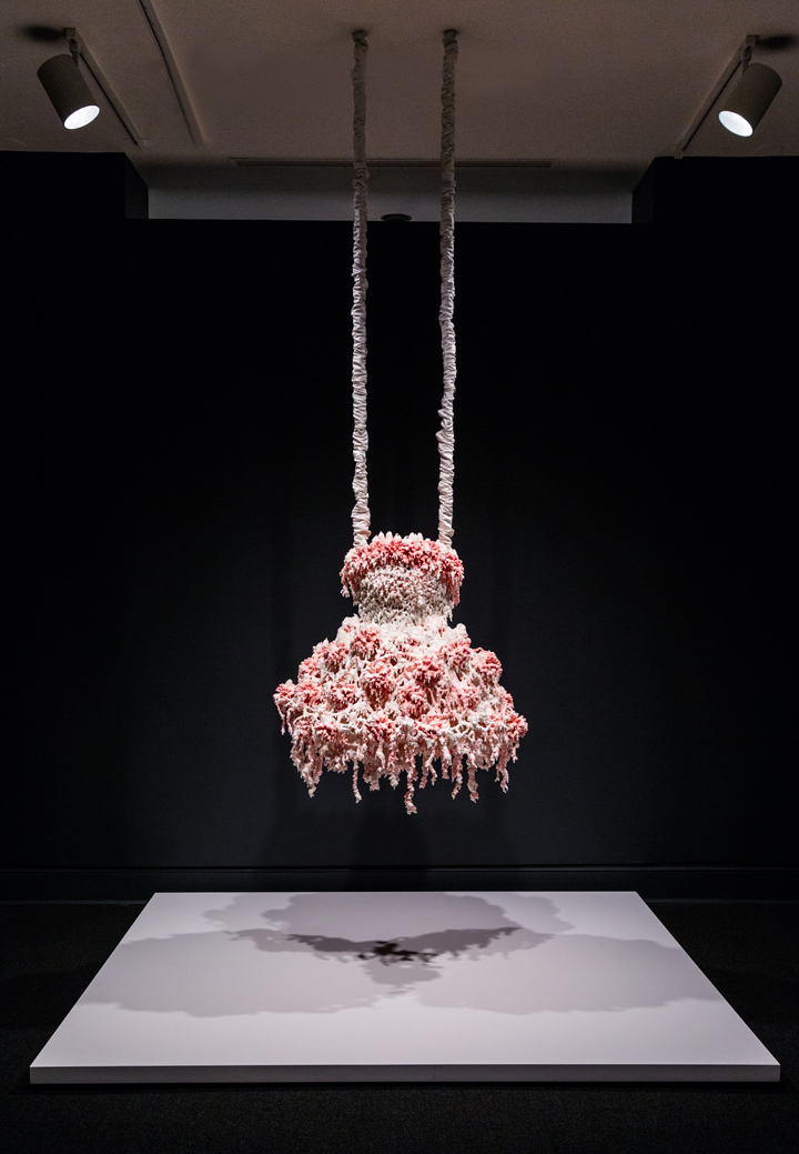 Installation view of a sculpture hanging from the ceiling in a gallery space. The sculpture consists of myriad layers of melted pink and white wax that encrust and obscure the metal armature for this abstract sculpture, which hangs from satin-wrapped chains. Its color and shape, as well as the bumpy, lacy texture, evoke a frilly tutu, lavishly frosted wedding cake, or coral accretions.