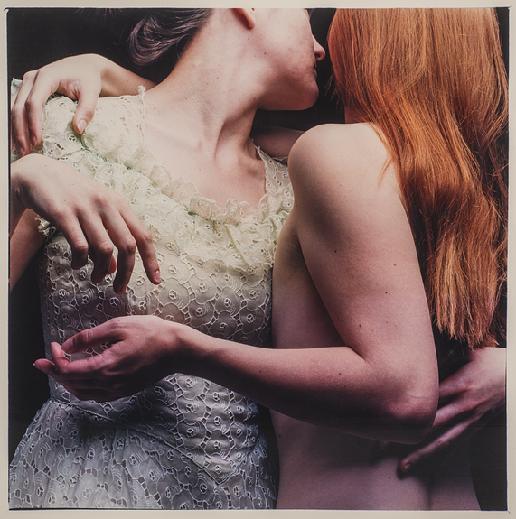 Two people, one clothed in a lace dress and the other nude, with their arms around each other and facing away from the viewer. The person on the left has brown hair pulled back and the person on the right has long, red hair.