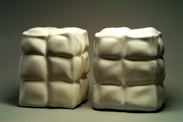 Two cubes in white sit next to each other. The sculptures look soft, as the light bounces off of them; yet, they also look like they are made out of a hard material  like stone. Several squares are cut out in them, making it look as if invisible threads were running through the sculptures. 